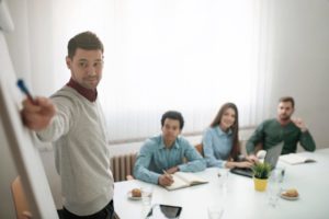 Un groupe de personnes en salle de réunion regarde un tech lead faire une démonstration au tableau.